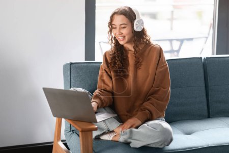 Photo for Teen girl wearing headphones watching movie on laptop, enjoying digital leisure and e-learning, sitting on sofa at home interior. Internet technologies and gadgets concept - Royalty Free Image