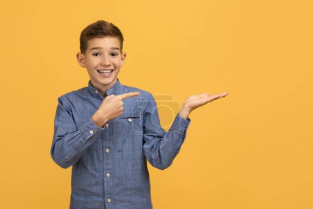 Foto de Cheerful teenage boy in denim shirt pointing at his open palm, happy male kid presenting an invisible product with beaming smile, standing isolated over yellow studio background, copy space - Imagen libre de derechos