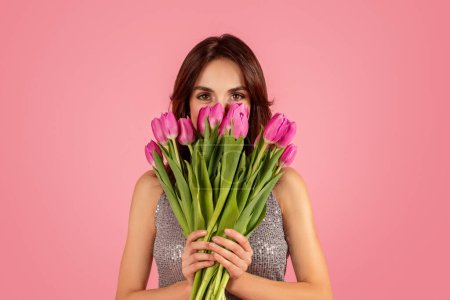 Téléchargez les photos : Enigmatic woman peering over a lush bouquet of pink tulips, her eyes captivating with a hint of mystery against a soft pink background. Spring holiday celebration event, ad and offer - en image libre de droit