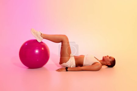 Photo for A relaxed latin woman with a shaved head lies on her back performing a bridge exercise with her feet on a pink fitness ball, in a serene environment with a gradient background - Royalty Free Image