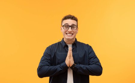 Photo for Pleading begging young man 20s wearing basic casual clothing and eyeglasses standing holding hands folded in praying gesture looking camera isolated on yellow color wall background, studio portrait - Royalty Free Image