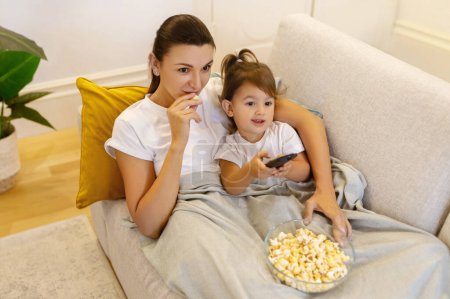 Photo for Mom and daughter watching TV with popcorn on sofa, relaxed young mother and preschooler female child lying under blanket, bonding together while resting in living room at home, - Royalty Free Image