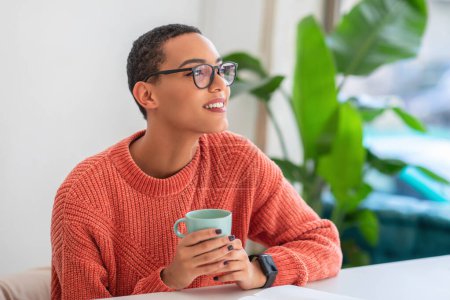 Photo for Joyful happy latin young woman with glasses wearing an orange sweater, holding a mug, and looking away with a smile in a well-lit room with lush plants enjoy spare time, free space - Royalty Free Image
