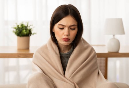 Photo for Closeup of upset beautiful brunette young woman wrapped in blanket sitting on couch, freezing at home, feeling cold during winter season in unheated apartment - Royalty Free Image