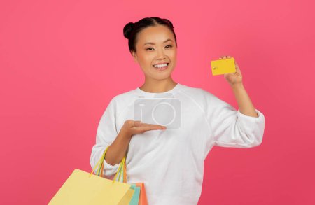 Téléchargez les photos : Cashback Concept. Happy Asian Woman Posing With Credit Card And Paper Shopping Bags Over Pink Background, Cheerful Korean Female Enjoying Big Discounts And Purchase Offers, Copy Space - en image libre de droit