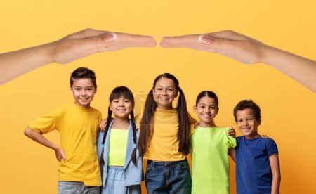 Téléchargez les photos : Mother love, care, protection. Group of cheerful beautiful multiethnic kids school aged boys and girls standing under woman hands roof, yellow background, collage - en image libre de droit