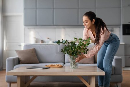 Foto de Smiling woman in peach blouse and blue jeans carefully arranges plant in vase on wooden coffee table in bright, modern living room setting - Imagen libre de derechos