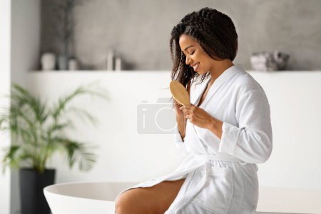 Téléchargez les photos : Haircare. Smiling Black Female Brushing Hair With Hairbrush, Happy African American Woman Wearing Bathrobe Detangling Split Ends While Sitting On Bathtub In Modern Bathroom At Home, Copy Space - en image libre de droit