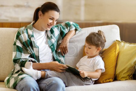 Foto de Cute little girl and her young mother reading book together at home, mom and preschool daughter sharing learning moment, relaxing on comfortable couch, fostering education and family bonds - Imagen libre de derechos