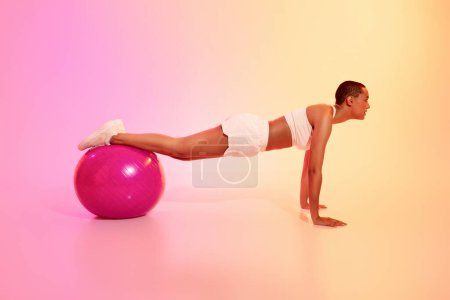 Téléchargez les photos : A disciplined woman with a shaved head holds a plank position on a pink exercise ball, demonstrating focus and strength in her white athletic wear against a gradient background - en image libre de droit