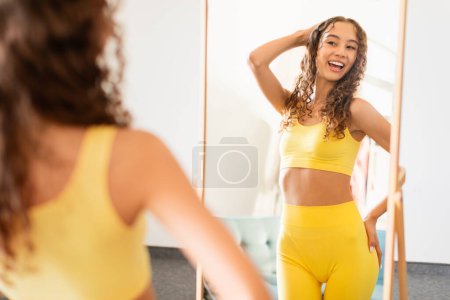 Téléchargez les photos : Confident young teen girl in sportswear, admiring her fit body smiling to mirror and her perfect reflection at home. Concept of self-love and fitness motivation. Selective focus - en image libre de droit