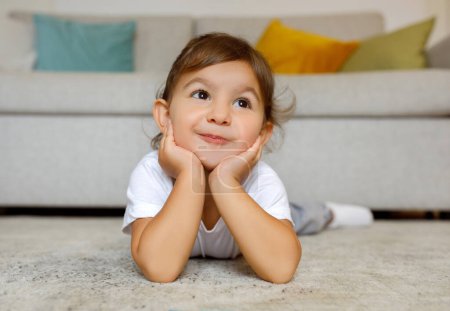 Foto de Adorable little girl with a charming smile lies on her stomach on the floor, hands on cheeks, cute toddler female child looking up with a dreamy, joyful expression in cozy home setting, copy space - Imagen libre de derechos