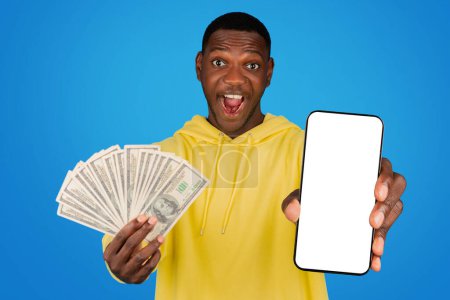 Foto de Excited young african american man in a yellow hoodie shows off a stack of money in one hand and a blank screen smartphone in the other against a blue studio background - Imagen libre de derechos