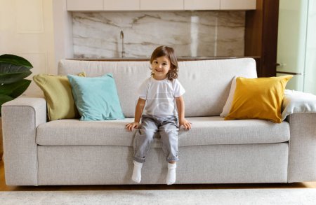 Téléchargez les photos : Cute preschool little girl sitting on couch at home, smiling at camera with innocence and joy, adorable female child embodying childhood happiness in comfortable living room interior, copy space - en image libre de droit