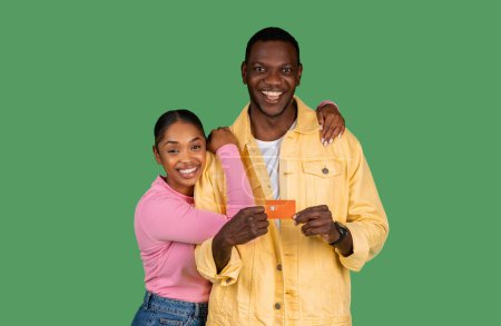 Téléchargez les photos : Happy african american couple holding credit card, recommending service and smiling at camera, embracing on green studio background. Bank customers showing card for payments - en image libre de droit