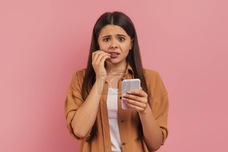 Téléchargez les photos : Anxious teenage girl biting her nails while looking at smartphone, concerned teen female standing against pink studio background, depicting stress or nervous anticipation, copy space - en image libre de droit