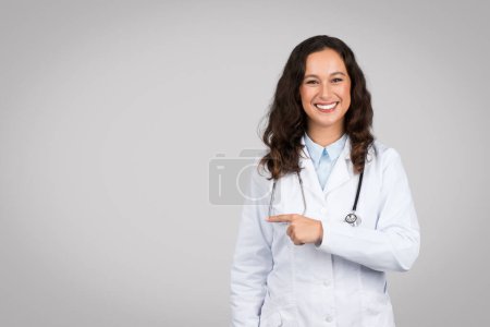 Téléchargez les photos : Cheerful young female doctor in white lab coat pointing to her side at free space with stethoscope around her neck against plain grey background - en image libre de droit