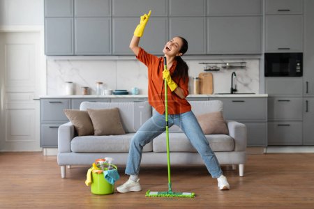 Photo for Playful woman in orange shirt and denim jeans, with yellow gloves, dances with green mop by sofa and bucket of cleaning tools, enjoying housework - Royalty Free Image