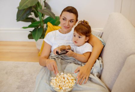 Photo for Mom and cute little daughter watching TV with popcorn on sofa, smiling family mother and preschooler female kid relaxing together at home, resting on comfy couch, child holding remote controller - Royalty Free Image