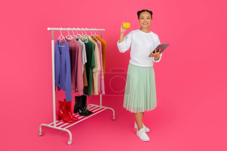 Foto de Asian woman holding credit card and digital tablet while standing near clothing rack, happy korean female browsing fashion choices, posing isolated on pink studio background, copy space - Imagen libre de derechos
