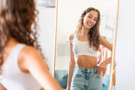 Photo for Weight Loss Concept. Happy Young Teen Age Girl Wearing Jeans After Dieting, Comparing Size Before And After Slimming, Posing Near Mirror Standing At Home. Selective Focus, Copy Space - Royalty Free Image