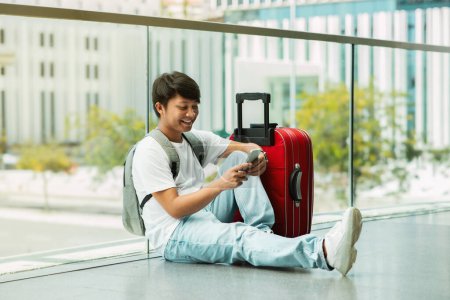 Photo for Cheerful young chinese man in comfy casual outfit going vacation alone, sitting on floor at airport with red luggage, using cell phone, online check-in, copy space, full length - Royalty Free Image
