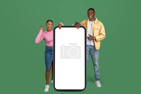 Téléchargez les photos : Yes. Excited Black Couple Pointing At Big Smartphone With Empty White Screen Shaking Clenched Fists, Cheerful Guy And Lady Celebrating Win, Standing On Green Background, Mock Up Collage, Copy Space - en image libre de droit