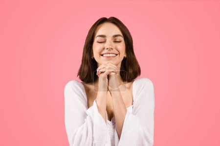 Photo for Content young caucasian woman with eyes closed, hands clasped in delight, radiates gratitude and serenity in a white blouse, embodying peaceful joy against a pink backdrop - Royalty Free Image