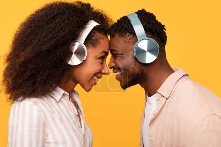 Téléchargez les photos : Intimate and joyful moment as young black couple in wireless headphones, touching foreheads and smiling, standing on yellow background, side view - en image libre de droit