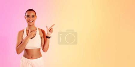 Photo for An engaging woman with a shaved head points to her left with a bright smile, dressed in a white sports bra and pale pink shorts against a soft gradient background, panorama - Royalty Free Image
