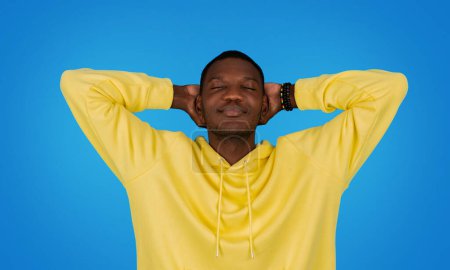 Téléchargez les photos : A serene african american man in a vibrant yellow hoodie with closed eyes, hands behind his head, and a tranquil expression, posing against a bright blue studio background - en image libre de droit