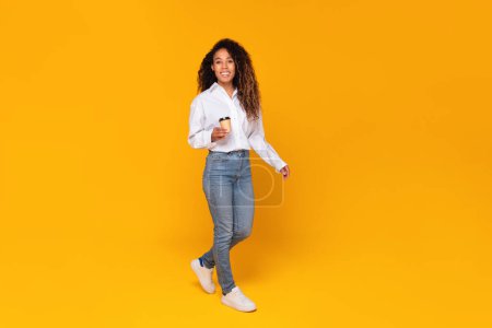 Photo for Full length shot of cheerful black lady holding paper coffee cup while walking and smiling to camera on yellow studio background, enjoying her break and tasty drink concept - Royalty Free Image