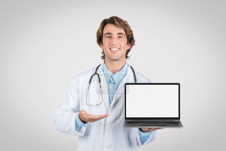 Téléchargez les photos : Cheerful male doctor presenting an open laptop with blank screen for mockups, ideal for medical software or telehealth services, against a grey backdrop - en image libre de droit
