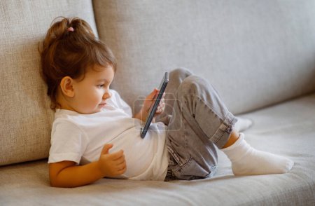 Foto de Cute toddler girl wearing white shirt using smartphone while sitting on cozy sofa, curious preschool female kid immersed in digital world, watching cartoons online or playing mobile games - Imagen libre de derechos