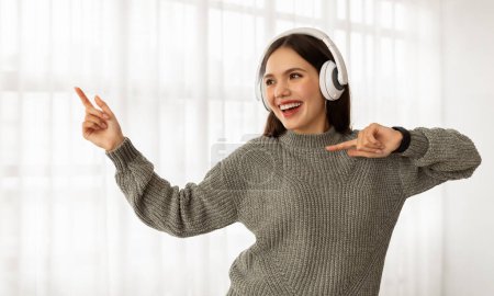 Foto de Joyful pretty brunette young woman listening to music and singing, dancing and smiling, using wireless headphones, home interior, copy space. Stress relief concept - Imagen libre de derechos