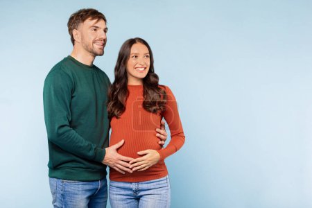 Foto de Expectant couple with the mans hands gently cradling the womans pregnant belly, both looking forward with happiness and anticipation, set against tranquil blue background, free space - Imagen libre de derechos