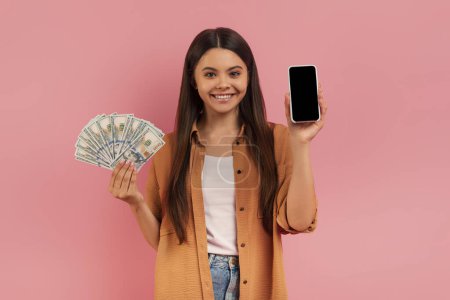 Photo for Happy Teen Girl Holding Blank Smartphone And Dollar Cash In Hands, Cheerful Female Teenager Posing Over Pink Studio Background, Illustrating Concept Of Mobile Payments Or Financial App, Mockup - Royalty Free Image