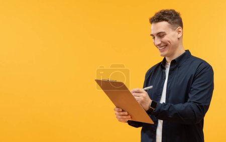 Téléchargez les photos : Happy handsome young man taking notes in clipboard, yellow studio background, copy space for advertisement. Guy worker employee writing down on board, filling form - en image libre de droit