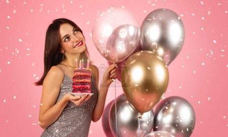 Téléchargez les photos : A beaming woman with dark hair, wearing a sequin silver dress, holds a vibrant stack of donuts with candles amidst a shower of confetti and metallic balloons on a pink background - en image libre de droit