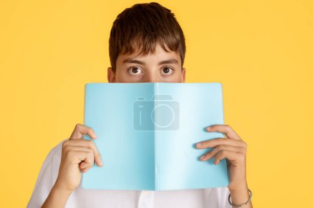 Téléchargez les photos : A boy with an intrigued expression carefully covers his face with a light blue book, leaving only his eyes visible, against a contrasting bright yellow background, close up - en image libre de droit