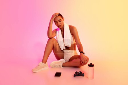 Foto de Tired sad latin young woman with a shaved head sitting on the floor, resting with a towel over her shoulder, a water bottle, and a phone next to her against a pink gradient - Imagen libre de derechos
