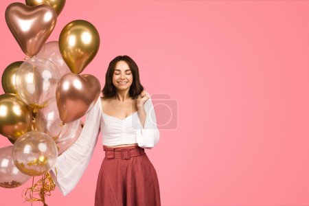 Foto de Contented caucasian glad young woman in a stylish white top and maroon skirt, holding a bunch of gold and clear balloons with a gentle smile, against a soft pink backdrop - Imagen libre de derechos