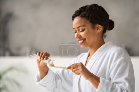 Foto de Oral Hygiene. Beautiful Black Woman Applying Toothpaste On Eco Bamboo Toothbrush At Home, Smiling Young African Woman Making Daily Dental Care, Standing In Bathroom Interior, Copy Space - Imagen libre de derechos