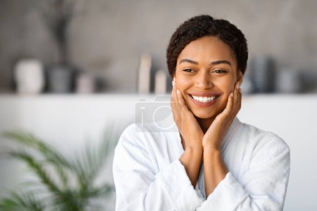 Photo for Beautiful Black Young Woman Wearing White Bathrobe Standing In Stylish Bathroom Interior, Happy African American Female Touching Face And Smiling, Enjoying Beauty Treatments And Skincare, Copy Space - Royalty Free Image