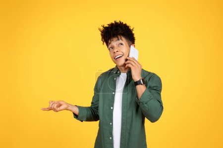 Téléchargez les photos : Talkative cheerful stylish handsome young black guy wearing casual clothing talking on phone with friend or lover and gesturing, sharing rumors, yellow studio background - en image libre de droit