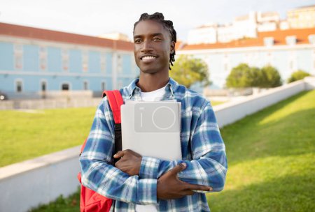 Photo for Casual black adult student guy with backpack, laptop and workbooks posing outside, over backdrop of university building and campus park. Education and knowledge concept - Royalty Free Image