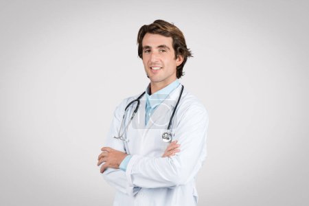 Téléchargez les photos : Smiling young european male doctor in white coat with stethoscope around his neck standing confidently with arms crossed against gray background - en image libre de droit
