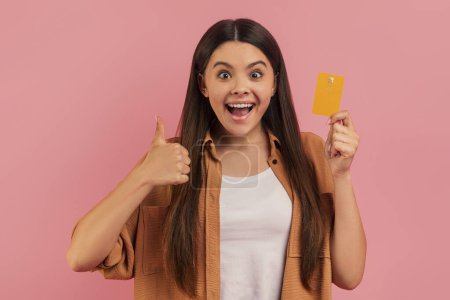 Téléchargez les photos : Ecstatic teenage girl holding credit card and giving thumb up, joyful female teen exuding excitement and approval, standing against pink studio background, symbolizing successful payment - en image libre de droit