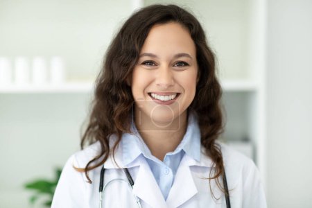 Photo for Closeup portrait of cheerful attractive brunette young european woman doctor wearing medical uniform white coat and stethoscope posing at clinic or hospital, smiling at camera - Royalty Free Image