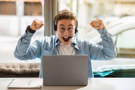 Photo for Glad young european guy in glasses, headphones using laptop, rises fists up, celebrate win, success at remote work or online communication in cafe. Winner gesture, game - Royalty Free Image
